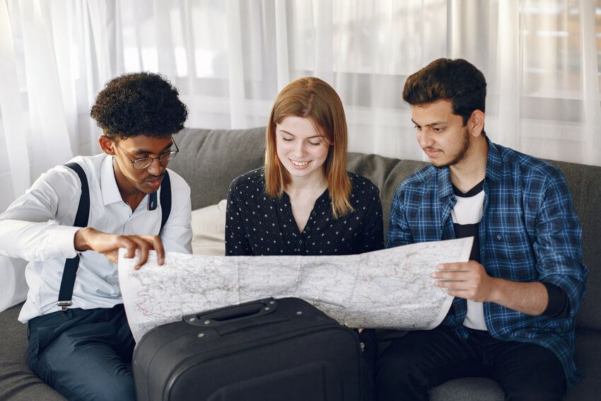 Dois homens jovens nas pontas de um sofá e uma mulher jovem no meio escolhendo num mapa o destino da viagem